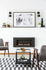 Poster above black fireplace in apartment interior with grey armchairs and patterned carpet. Real photo