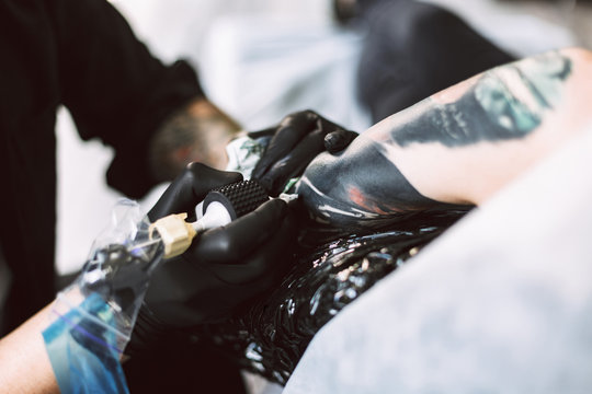 Close up professional tattooer in black gloves doing tattoo on hand using tattoo machine in studio