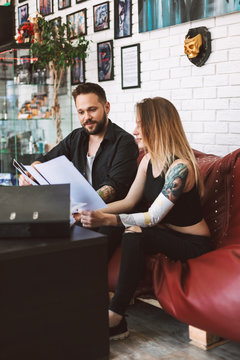 Professional tattoo master sitting on sofa with girl discussing sketch of new tattoo in tattoo studio