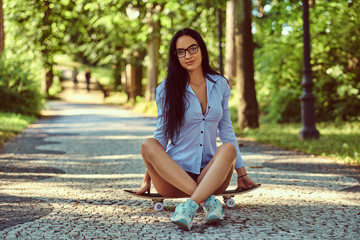 Gorgeous sensual brunette girl in glasses wearing a shirt and shorts sitting on a skateboard in a...