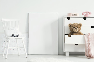 Teddy bear and pink blanket in cabinet next to empty poster with mockup in baby's room interior....