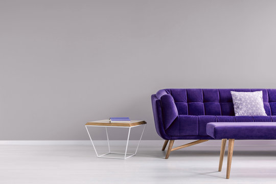 Pillow on a stylish, vibrant purple settee and a diamond shape, side table in a gray living room interior with place for a floor lamp. Real photo.