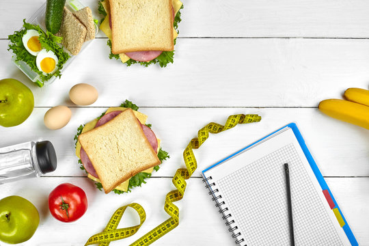 Top view planning notebook with copy space and healthy breakfast. Sandwich with ham and cheese, eggs, cucumber, tomato and bottle of water on white wooden table.