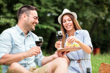 Happy young couple enjoying a glass of wine on a romantic picnic in a park. Man offering fruits and snacks on a wooden plate to his girlfriend..Love and tenderness, dating, romance, lifestyle concept