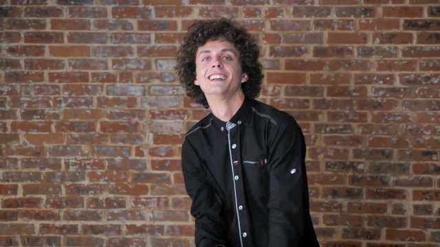 Young handsome cheerful man with curly hair dancing and looking at camera, brick wall background