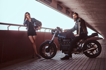 Obraz na płótnie Canvas Portrait of attractive couple - brutal bearded biker in black leather jacket sitting on a motorcycle and his young sensual brunette girlfriend on a footway under a bridge, looking at a camera.
