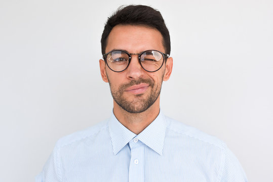 Portrait of unshaven handsome male frowns, being dissatisfied with something, blink with eye, wears round glasses and blue shirt, isolated over white studio background. People and emotion concept.