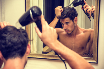 Shirtless young man drying hair with hairdryer, looking at himself in mirror at home