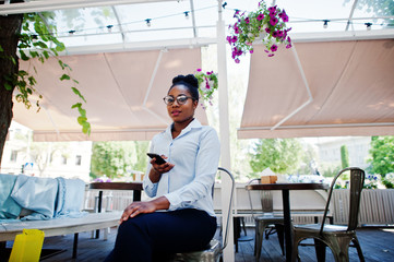 African american girl wear in glasses with mobile phone sitting at outdoor caffe.