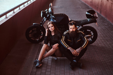 Fototapeta na wymiar Brutal bearded biker in black leather jacket and sensual brunette girl sitting together on a skateboard near custom-made retro motorcycle on a footway under a bridge.