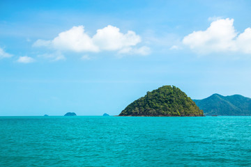 Small green island of pyramid shape in the tropical sea against a blue sky with white clouds. In the background mainland.