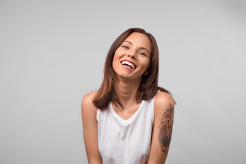 Smiling positive female with attractive look, wearing loose white T-shirt, posing against white blank wall. Happy woman with dark hair showing positive emotions after receiving pleasant compliment