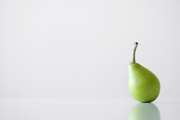 One green yellow pear with a twig stands on a white table, a reflection, a bright light, minimalism and free space for text.