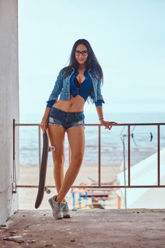 Seductive hot brunette girl wearing short shorts and jeans jacket holds a skateboard while standing to lean on a guardrail against the sea coast.