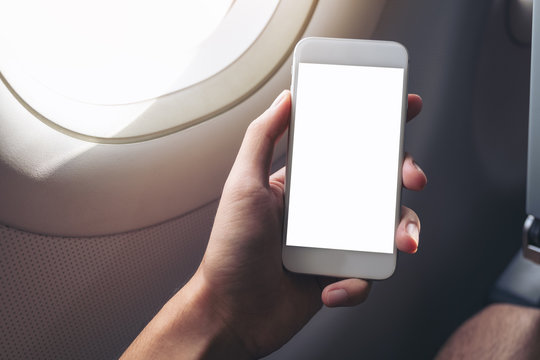Mockup image of a hand holding a white smart phone with blank desktop screen next to an airplane window