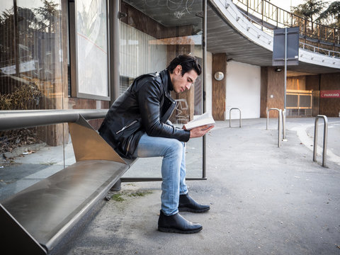 Side view of handsome stylish man in leather jacket sitting on bench on street and reading book with smile