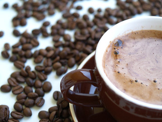 eine braune Tasse mit Cappuccino, daneben liegen Kaffeebohnen