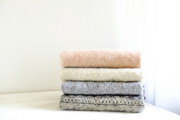 Fototapeta na wymiar Bunch of knitted warm pastel color sweaters with different knitting patterns folded in stack on white wooden table, textured wall background. Fall winter season knitwear. Close up, copy space for text