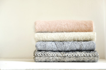 Bunch of knitted warm pastel color sweaters with different knitting patterns folded in stack on white wooden table, textured wall background. Fall winter season knitwear. Close up, copy space for text