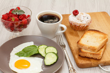 Plate with fried egg, cut cucumber and leaf of basil, glass bowl with raspberries, cup of coffee, fork, muffin and toasts on wooden cutting board.