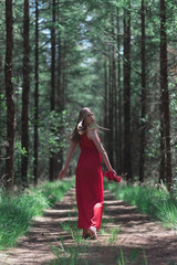 Blonde woman in red dress holding shoes in hand looking over shoulder. Walking on forest path.