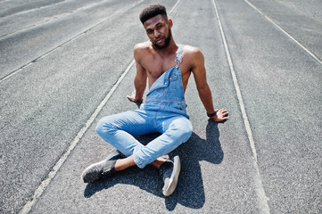 Handsome sexy african american bare torso man at jeans overalls sitting at stadium racecourse. Fashionable black man portrait.