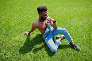 Handsome sexy african american bare torso man at jeans overalls sitting at green grass of stadium...