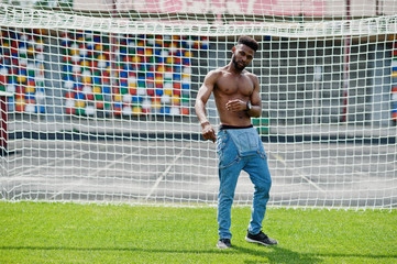 Handsome sexy african american muscular bare torso man at jeans overalls posed at green grass agasinst football gates of stadium field. Fashionable black man portrait.