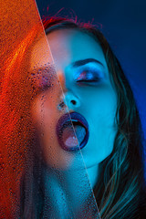 A beautiful model girl, illuminated with blue and red lights, half peeks out of a glass covered...