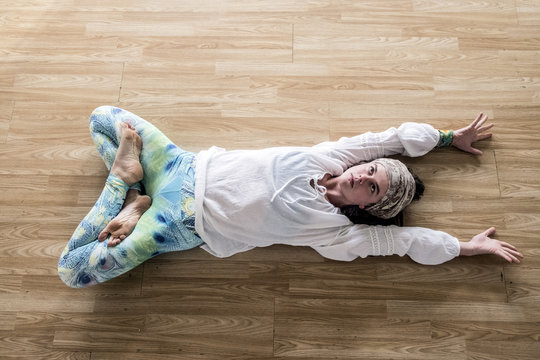 High angle view of young woman wearing headscarf and white blouse lying on her back on hardwood floor, doing yoga.