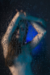 Fototapeta na wymiar Blurred silhouette of a beautiful slender naked girl standing behind glass covered with drops of water. Artistic and conceptual photo in blue tones