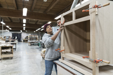 A Black woman carpenter working on a cabinet project in a large woodworking shop.