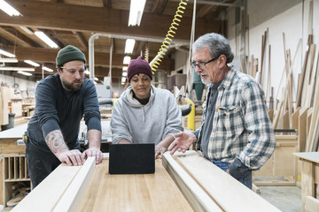 A group of mixed race carpenters discussing a project at a work station in a large woodworking shop.