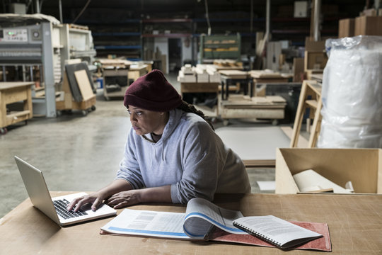 A Black female carpenter working on a lap top computer after work hours in a large woodworking factory.