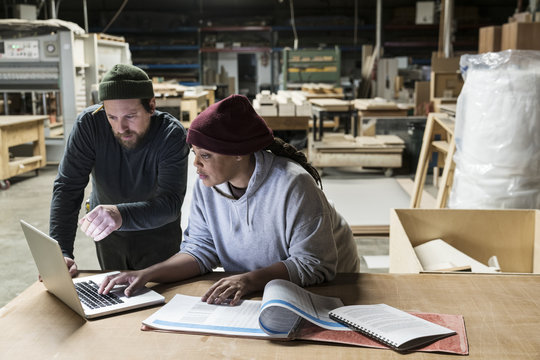 A Caucasian male carpenter and a Black female carpenter working on a laptop computer after work hours in a large woodworking factory.