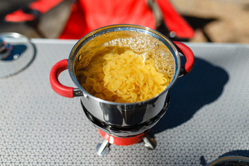 Closeup cooking in aluminum pan will boil with pasta on fire from the portable gas burner next to the plates and mugs on the portable table against the in the camping. Cooking traveling concept.