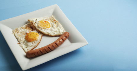 Traditional Clasic Breakfast Fried Eggs and Sausage on a white Square Porcelain Plate. Isoalted on Blue Background. Banner Copy Space.