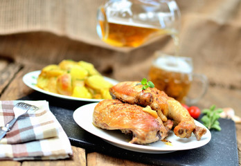 Golden grilled chicken legs on a plate, roasted potatoes and pouring cold beer into a glass, rosemary, tomatoes, oregano, salt, pepper and garlic on a wooden table - limited DOF