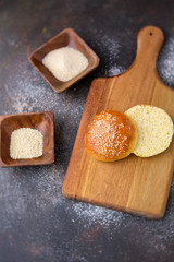 Homemade Hamburger Bun Topped with Sesame Seeds Cut Open on Wooden Cutting Board; Sesame Seeds and Sugar in Small Wooden Bowls