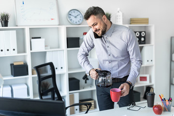 A young man in the office is standing near the table, holding the phone with his shoulder, looking at the monitor and pouring coffee into the cup.