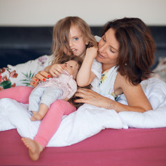 Beautiful happy family scene with mother talking to her little daughter with her doll, lying in bed at home