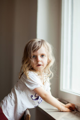 curious cute little girl standing on a chair at the window, blonde curls, looking into camera