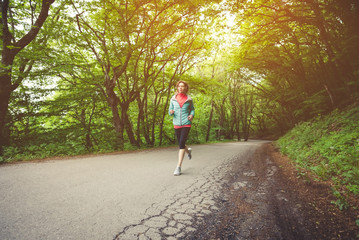 Young fitness blonde woman in headphones running at morning caucasian forest trail in sun light