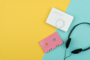 top view of a pink audio cassette tape with a white portable cassette player and a black on-ear headphone on the yellow and blue background