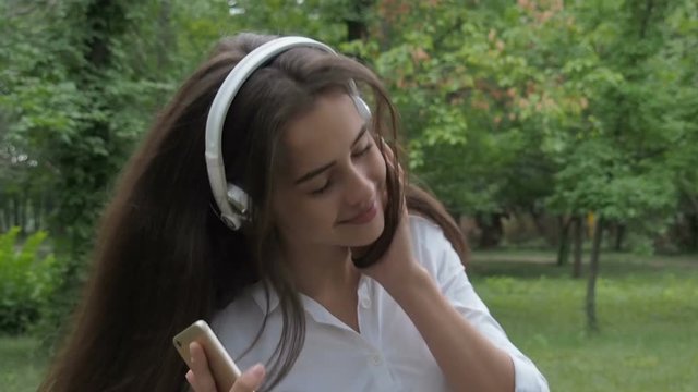 The girl dances in the headphones. A girl with a phone is dancing in the park.