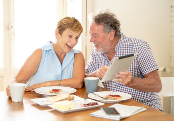 Beautiful senior retired old couple using digital tablet with joy while having breakfast at home