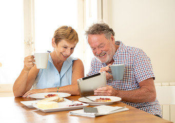 Beautiful senior retired old couple using digital tablet with joy while having breakfast at home