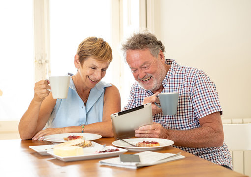 Beautiful senior retired old couple using digital tablet with joy while having breakfast at home