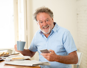 Handsome senior retired old man using mobile phone with joy while having breakfast at home
