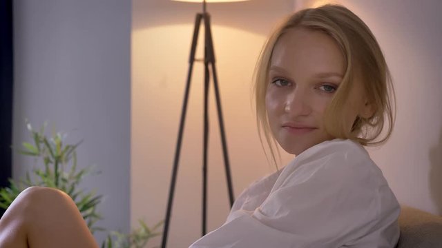 Young sexy blonda lady in lingerie and shirt is sitting on sofa, watching at camera, smiling, relax concept